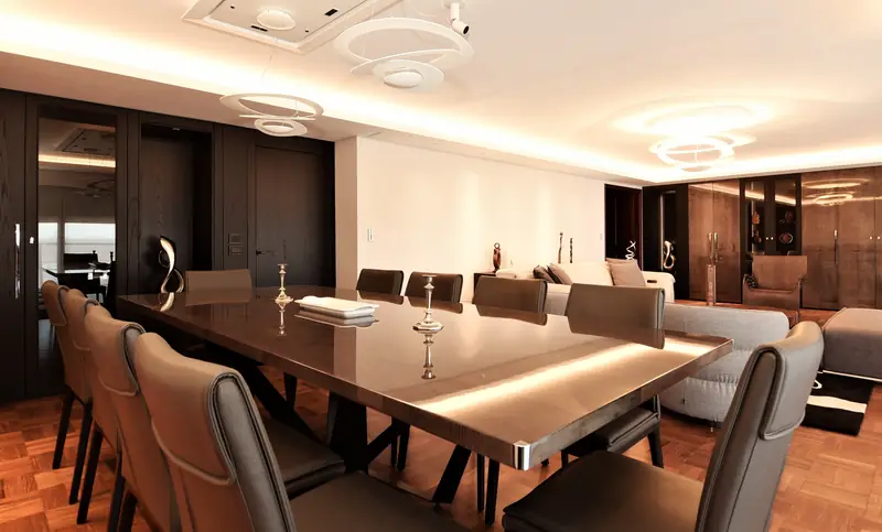 Comedor con mesa larga de madera lacada iluminada por debajo y sillas de cuero, bajo lámparas de anillos