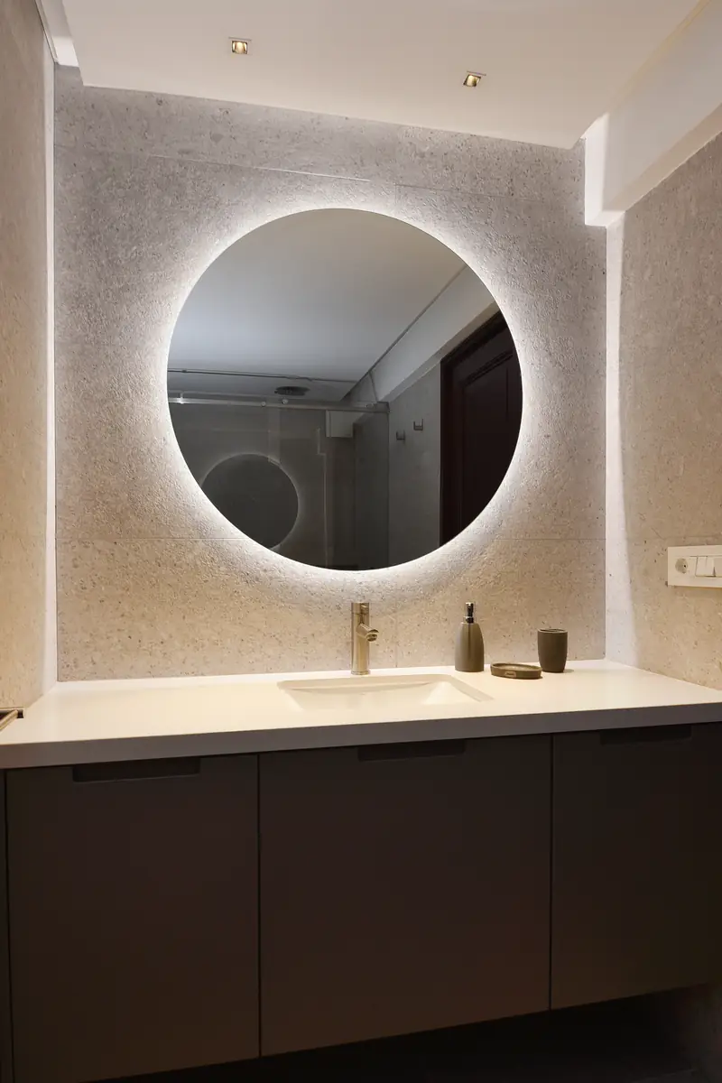 Espejo redondo retroiluminado sobre una pared de piedra clara y un mueble de baño oscuro