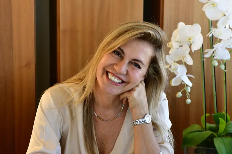 La arquitecta Karin Bia sentada en su escritorio, sonriendo, con orquídeas blancas y revistas de arquitectura