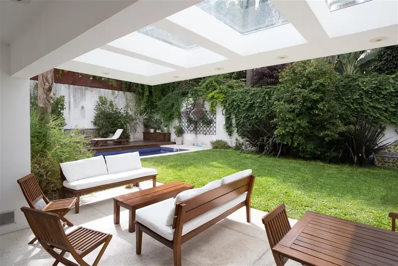Galería bajo lucernarios con sillones de madera y almohadones blancos, abierta al jardín con la piscina y un muro cubierto de enredaderas