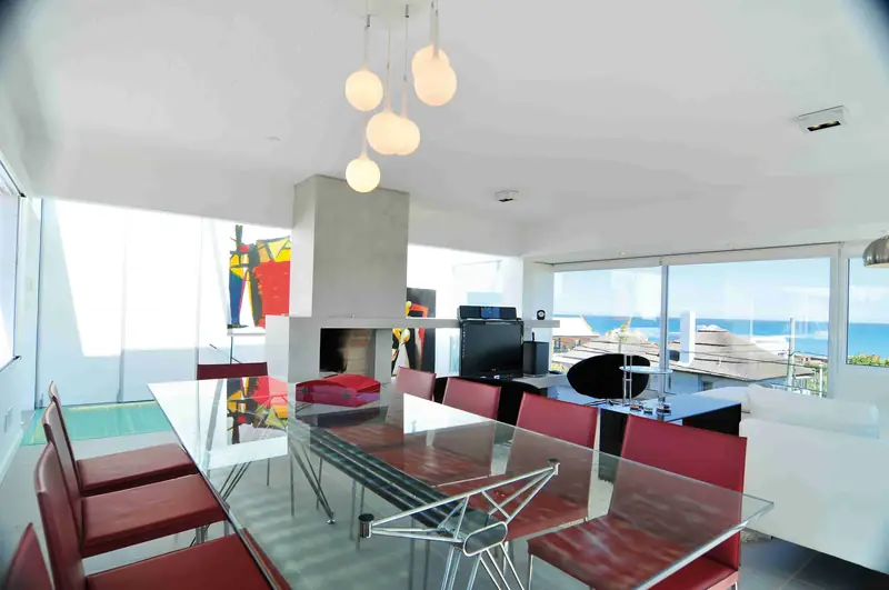 Comedor con mesa de cristal y sillas rojas bajo lámparas de globos blancos, junto a la chimenea gris y ventanales con vista al mar
