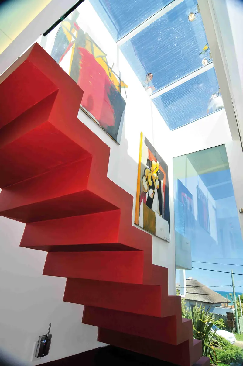 Escalera roja de peldaños volados vista desde abajo, junto a un muro blanco con cuadros y bajo un lucernario de vidrio
