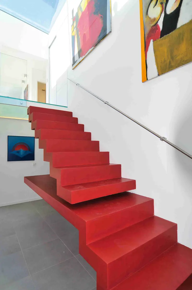 Escalera de peldaños rojos volados desde un muro blanco, con pasamanos de acero y cuadros de colores en la pared