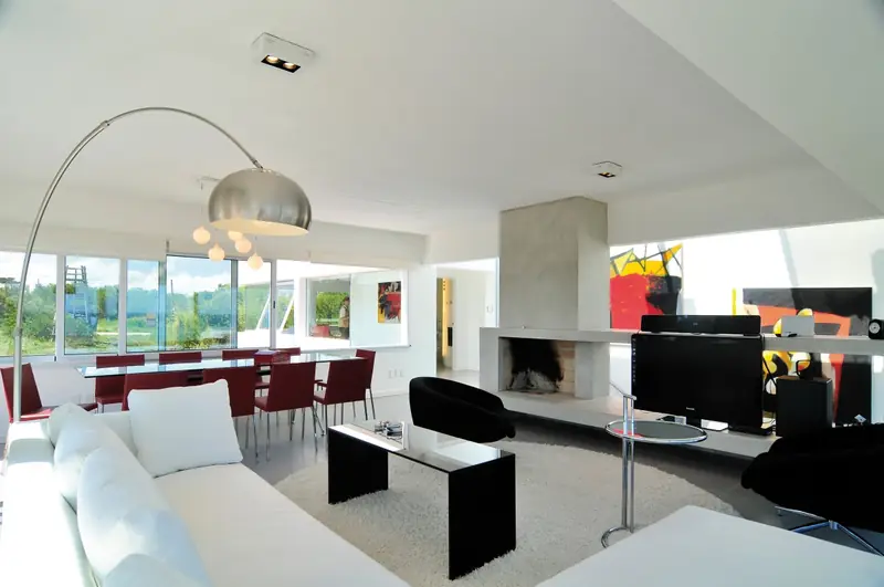 Estar y comedor continuos con sofá blanco, lámpara de arco, chimenea de hormigón exenta y ventanales a lo largo de toda la casa