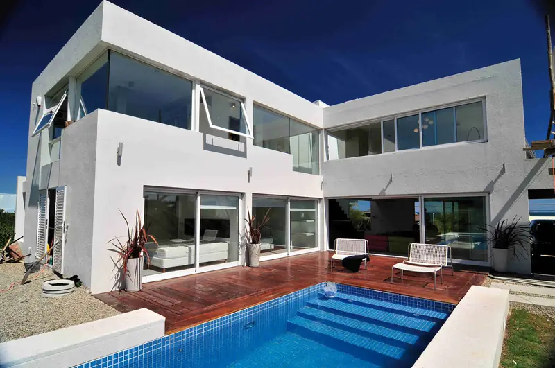 Fachada posterior de la casa: dos volúmenes blancos en L con grandes ventanales, un deck de madera y la piscina con escalones bajo un cielo azul intenso