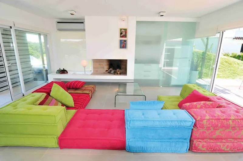 Sala de juegos con un sofá modular de cojines verdes, fucsias y celestes frente a una estufa a leña de ladrillo, entre ventanales