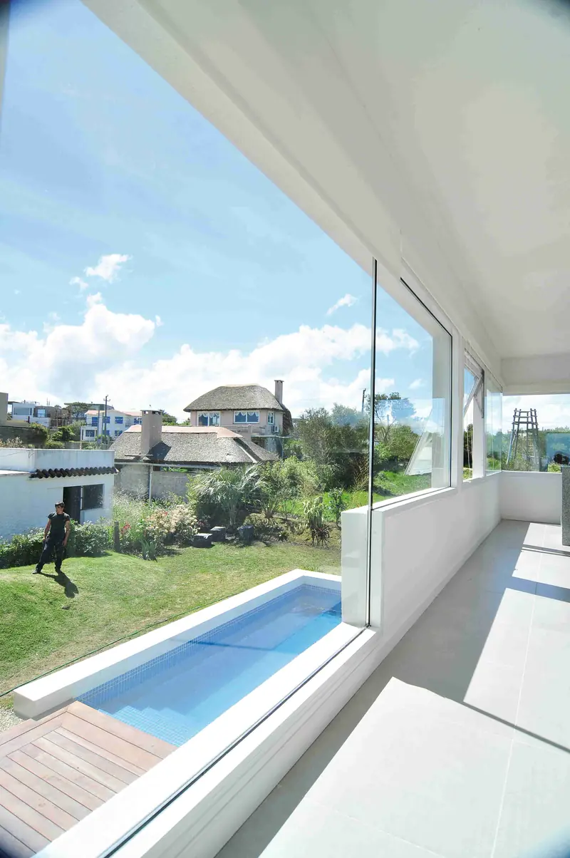 Pasillo junto a un ventanal de esquina que mira al jardín y a la piscina larga, con casas y cielo con nubes al fondo