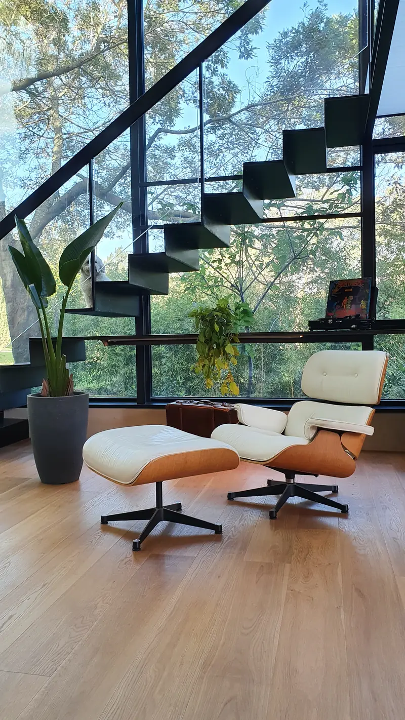 Sillón Eames de cuero blanco bajo la escalera flotante de peldaños negros, frente al muro de cristal y los árboles