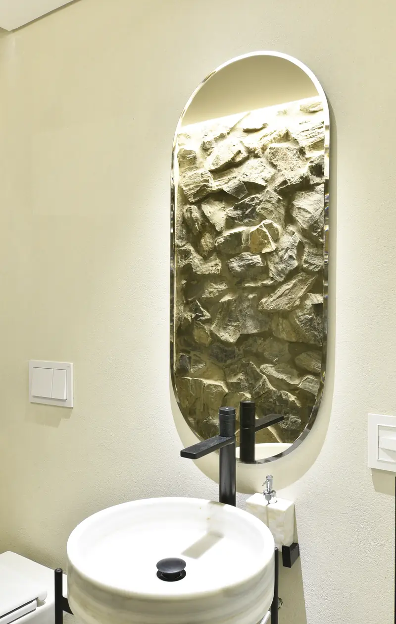 Baño con espejo oval iluminado que refleja un muro de piedra, sobre una bacha blanca redonda con grifería negra