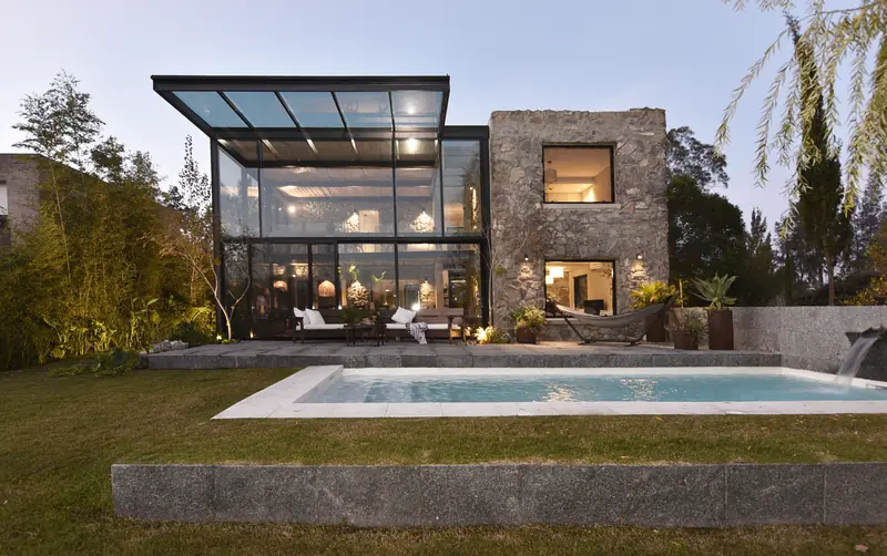 La casa al anochecer desde el jardín: caja de cristal iluminada, volumen de piedra y piscina sobre el césped