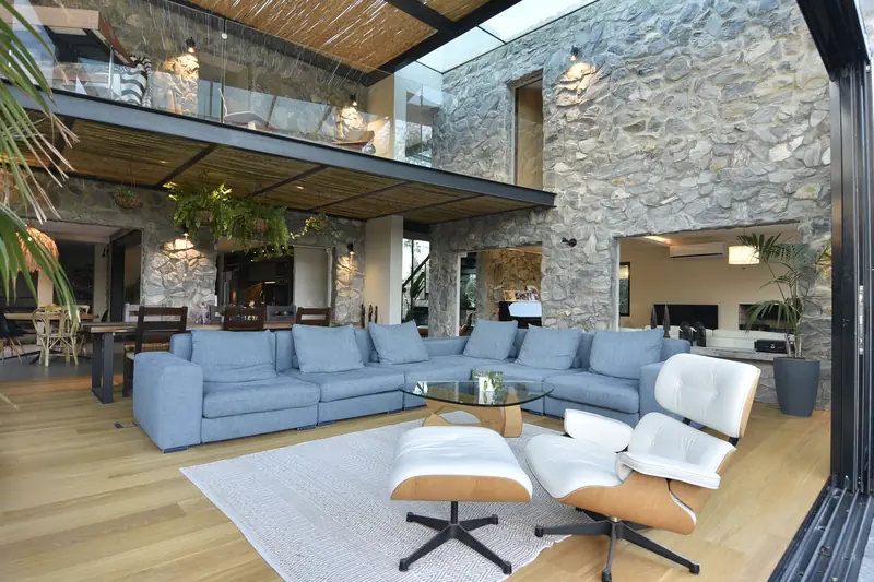 Galería bajo la pérgola con sofá azul y sillón Eames, ante la fachada de piedra