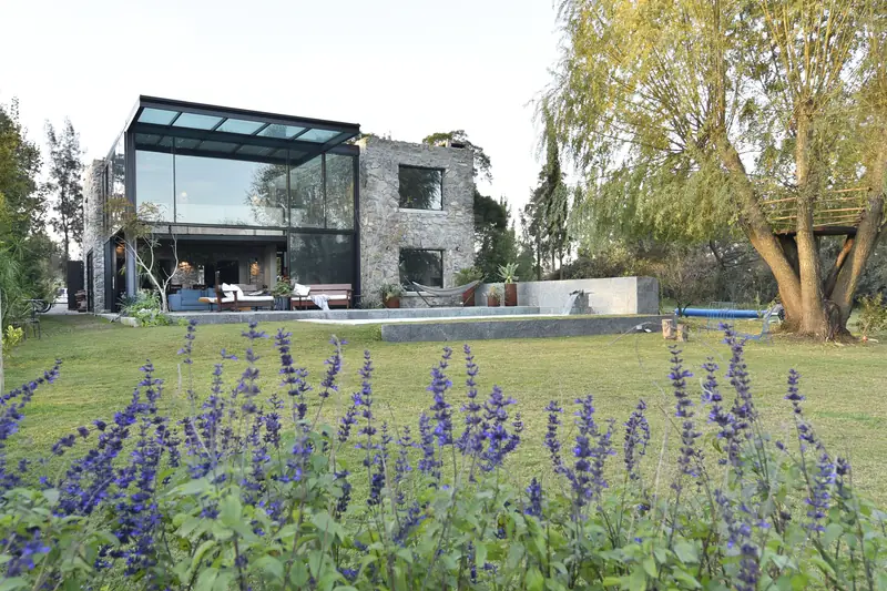 La casa de piedra y cristal al fondo del jardín, con flores azules en primer plano, la piscina y un sauce