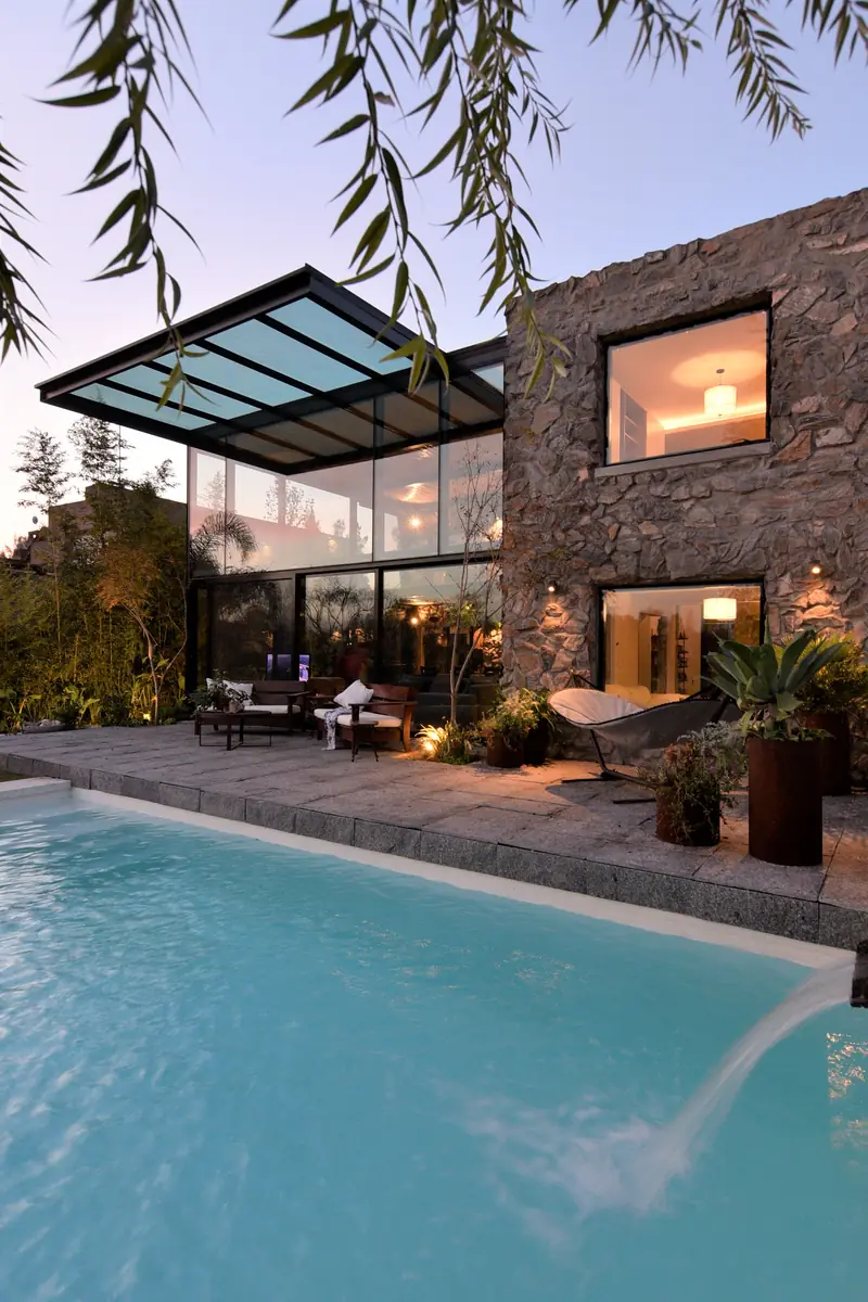 Piscina al anochecer frente a la caja de cristal iluminada y el volumen de piedra, bajo las ramas de un sauce