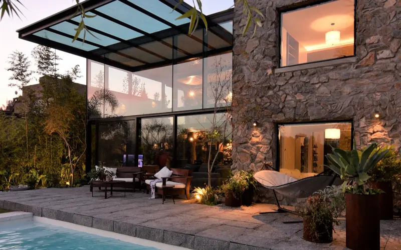 Piscina al anochecer frente a la caja de cristal iluminada y el volumen de piedra, bajo las ramas de un sauce