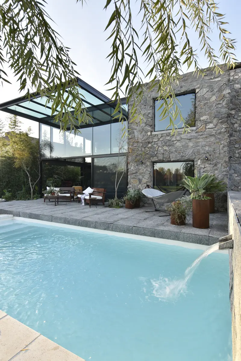 Piscina con caño de agua junto a la terraza de piedra y el volumen de piedra, bajo las ramas del sauce