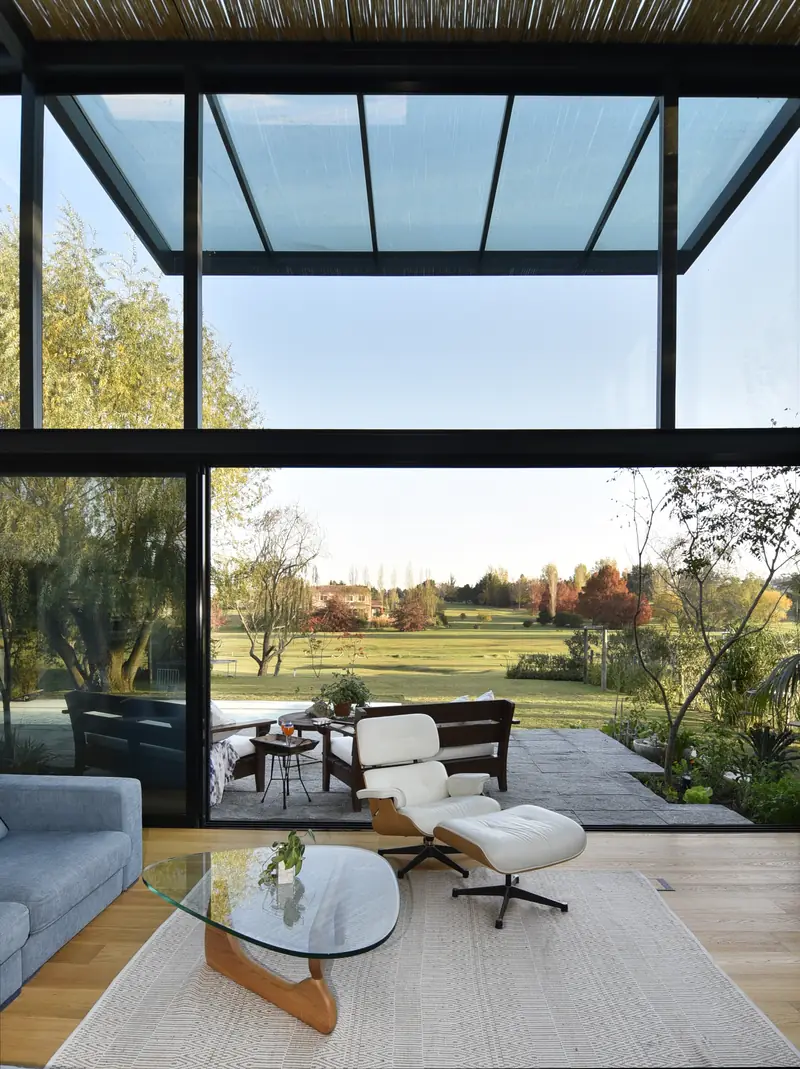 Sillón Eames y mesa de cristal junto al ventanal abierto al campo de golf, bajo la pérgola de vidrio
