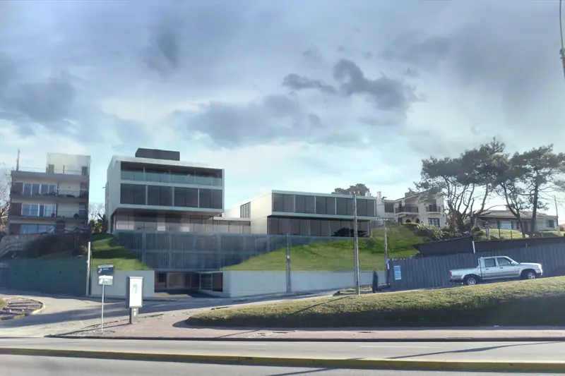 Edificios bajos de líneas horizontales sobre la loma verde de la rambla, bajo un cielo con nubes