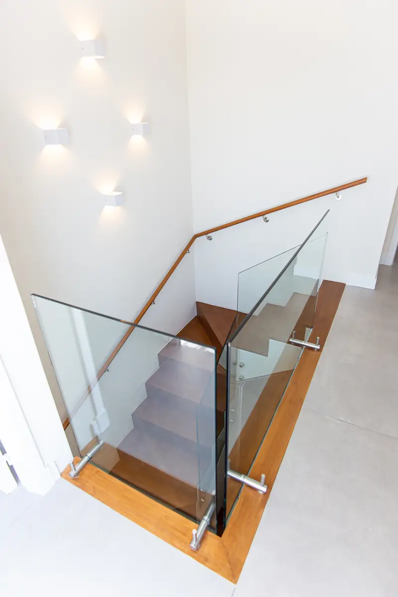 Escalera de madera con baranda de vidrio vista desde arriba, iluminada por apliques de pared