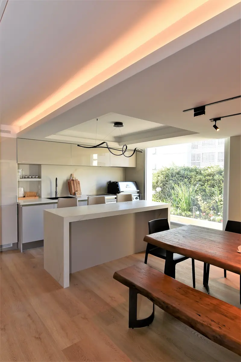 Cocina con isla blanca y mesa de madera con bancos junto a un ventanal hacia el jardín