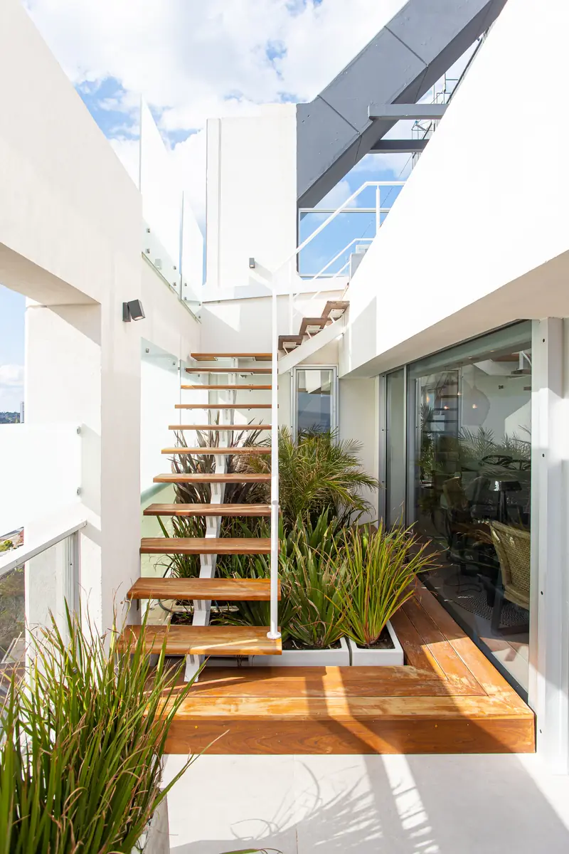 Escalera de peldaños de madera volados junto a un patio con plantas, que une los dos niveles del penthouse