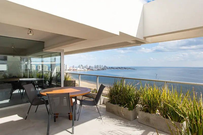 Terraza del penthouse con mesa y sillas junto a jardineras, frente al agua y la línea de la costa